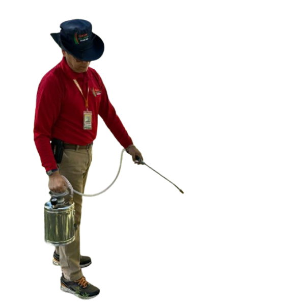 A person wearing a red shirt, khaki pants, and a dark hat is holding a sprayer with a long nozzle. They appear to be performing pest control or spraying outdoors.