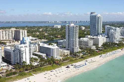 Aerial view of a beachfront cityscape with tall buildings and a sandy beach. The turquoise ocean stretches along the shore, with beachgoers, umbrellas, and greenery in the foreground. A calm bay and scattered clouds are visible in the background.